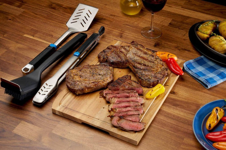 Stainless steel grilling tool set with spatula, tongs, and brush alongside grilled steaks on a wooden cutting board.